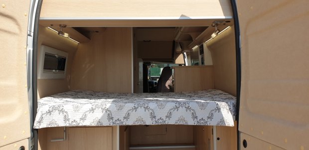 Wnętrze kampera z podwójnym łóżkiem, drewniane wykończenie i przedni fotel kierowcy.