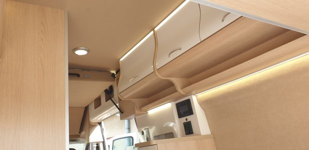 Wnętrze kampera z jasnymi drewnianymi szafkami nad głową i podświetleniem LED