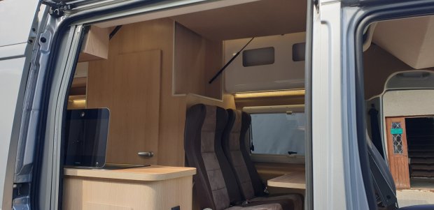 Wnętrze kampera: drewniana zabudowa, dwa ciemne fotele i stolik przy oknie