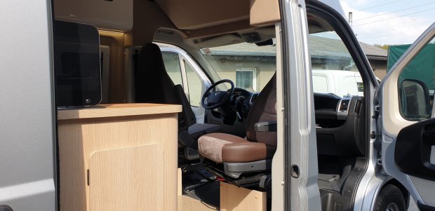 Wnętrze kampera z obróconym fotelem kierowcy i drewnianą szafką przy drzwiach.
