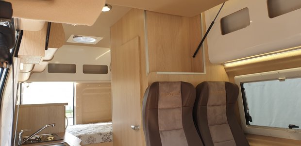 Wnętrze kamperu: drewniane meble, dwa brązowe fotele, aneks z kuchenką i tylnym łóżkiem