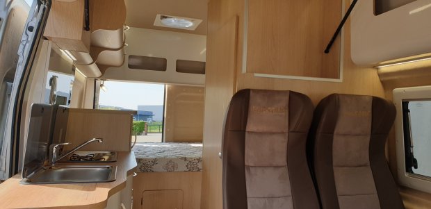 Wnętrze kampera: aneks kuchenny z umywalką i kuchenką, łóżko i dwa brązowe fotele.