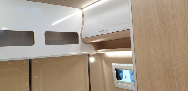 Wnętrze kampera: białe półki z drewnianymi wykończeniami, lampka i małe okno.