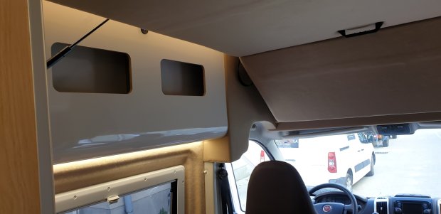 Przednia część kampera z półkami nad głową, fotelem kierowcy i widokiem na zewnątrz.