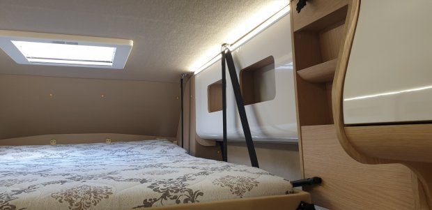 Wnętrze kampera: łóżko z wzorzystym materacem, świetlik i szafki z pasami.