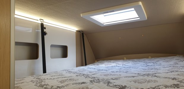 Górne łóżko w kamperze z oknem dachowym, paskami zabezpieczającymi i wzorzystą pościelą