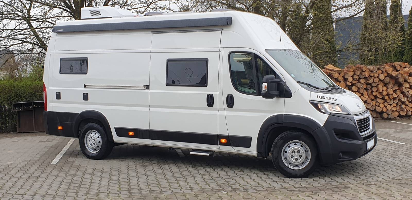 Biały kamper zaparkowany na bruku obok stosu polan drewna.