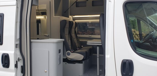 Wnętrze kampera z otwartymi drzwiami, dwiema fotelami przy stoliku i szafką