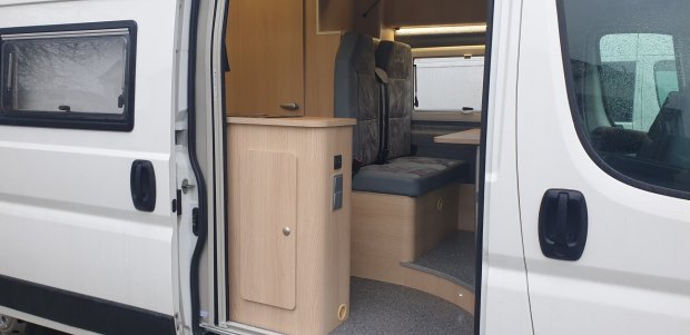 Wnętrze kampera przez otwarte drzwi: drewniana zabudowa i tapicerowane siedziska