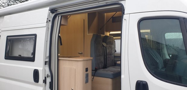 Wnętrze kampera przez otwarte boczne drzwi: aneks kuchenny i siedzenia.