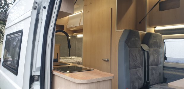 Wnętrze kampera z drewnianą zabudową, zlewozmywakiem, czarną baterią i dwoma szarymi siedzeniami.