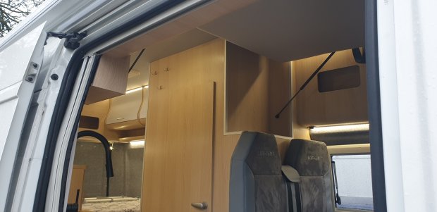 Wnętrze kampera z jasnym drewnem, szafkami, dwoma fotelami i łóżkiem.