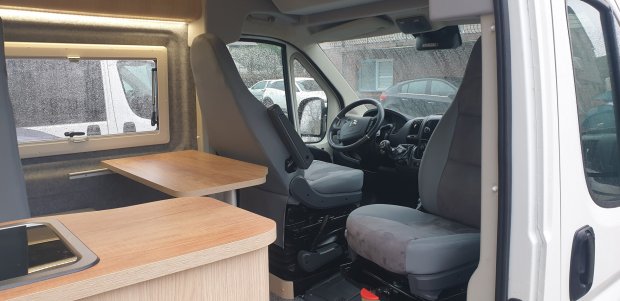 Wnętrze kampera: przednie fotele obrotowe, drewniany stolik i okno z kroplami deszczu