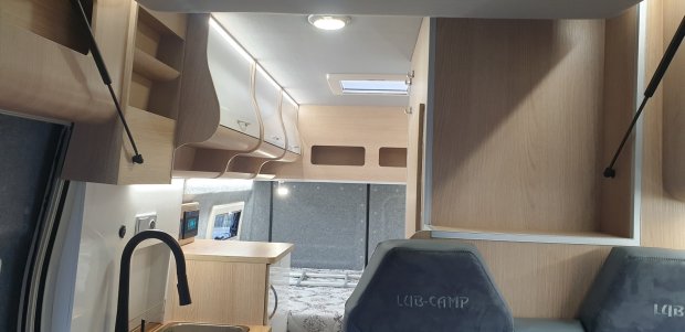 Wnętrze kampera: jasne drewniane szafki, zlew, dwa fotele i łóżko.