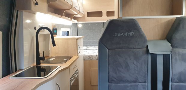 Wnętrze kampera z drewnianą kuchnią, czarnym kranem i szarymi fotelami LUB-CAMP