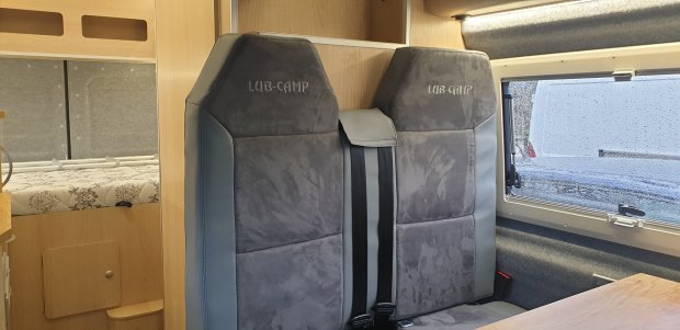 Dwa szare fotele kempingowe przy stole i oknie, w tle łóżko i szafka.