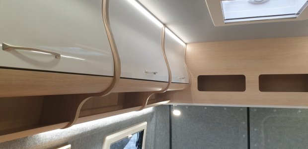 Wnętrze kampera z jasnymi drewnianymi półkami i białymi, błyszczącymi szafkami