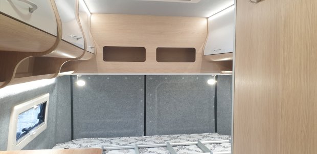 Wnętrze kampera: łóżko pod drewnianymi półkami, czytelnicze lampki i okno.