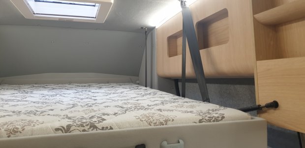 Łóżko w kamperze z jasnym materacem, drewniane schowki i świetlik nad głową