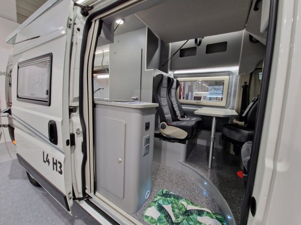 Wnętrze kampera z otwartymi drzwiami, stołem, dwoma fotelami i zabudową kuchenną.