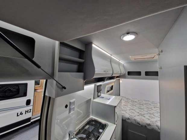 Wnętrze kampera z aneksem kuchennym, szafkami sufitowymi i łóżkiem.
