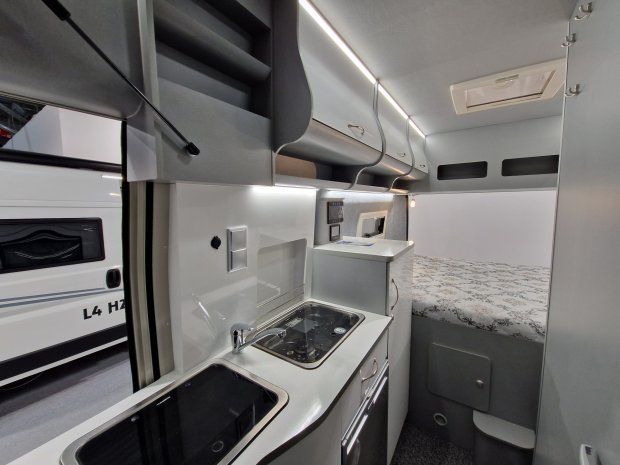 Wnętrze kampera: kuchnia ze zlewem i płytą, półki sufitowe i łóżko.