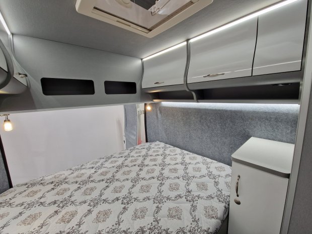 Wnętrze przyczepy/kampera: łóżko z wzorzystą pościelą, górne szafki i lampki.