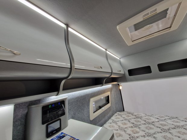 Wnętrze przyczepy kempingowej: półki sufitowe z oświetleniem, okno, panel sterowania i łóżko.