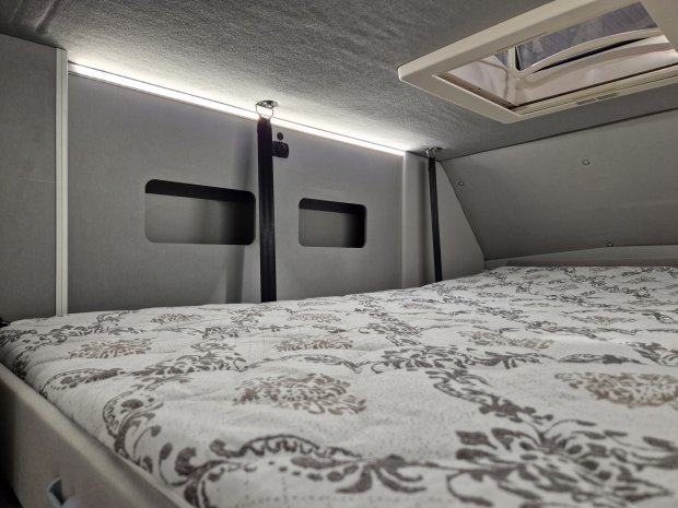 Wnętrze furgonu kempingowego: łóżko z wzorzystym materacem, okno dachowe i oświetlenie LED.