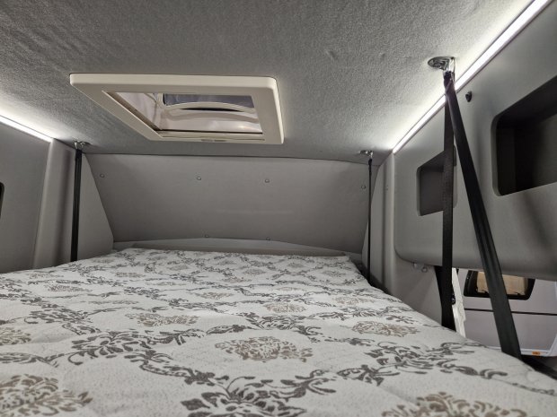 Wnętrze kampera: łóżko z wzorzystą pościelą, świetlna listwa i okno dachowe.