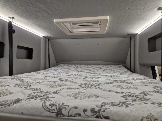 Wnętrze kampera: łóżko z wzorzystym materacem, szare ściany i dachowy właz