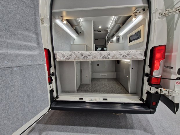 Tył kampera z uniesionym łóżkiem, schowkami i oświetlonym szarym wnętrzem