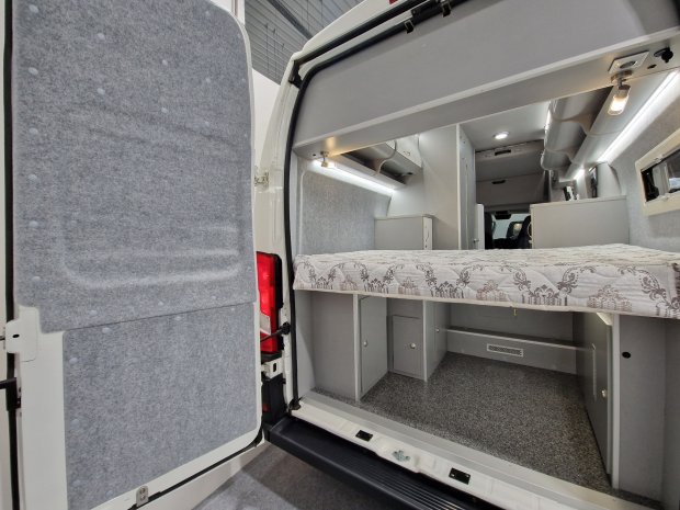 Wnętrze kampera widziane od tyłu z łóżkiem nad schowkami i szarymi szafkami.