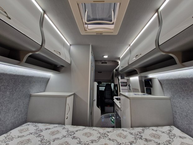 Wnętrze jasnego kampera z łóżkiem, szafkami i oświetleniem sufitowym