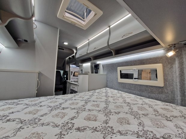 Wnętrze kampera z łóżkiem, górnymi szafkami i małym oknem