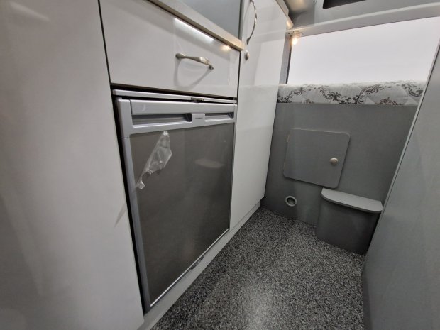 Wnętrze kampera: mała lodówka ze srebrnymi drzwiami, białe szafki i materac