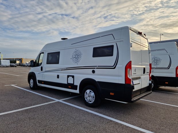 Biały kamper z kompasem na boku, zaparkowany na parkingu pod rozproszonymi chmurami