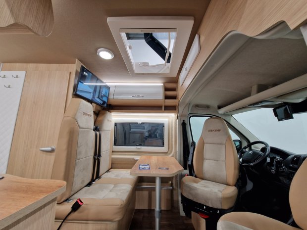 Wnętrze kampera z beżowymi siedzeniami, stołem, telewizorem i oknem dachowym