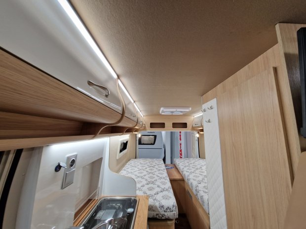 Wnętrze kampera z dwiema łóżkami, drewnianymi szafkami i kuchnią.