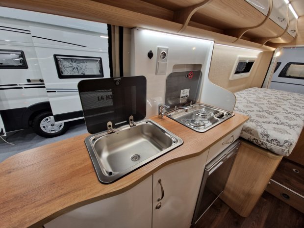 Mała kuchnia w kamperze: zlew, dwupalnikowa kuchenka i drewniany blat obok łóżka