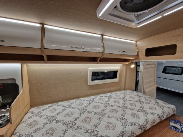 Wnętrze kampera: łóżko z wzorzystym materacem, górne szafki, okienko i oświetlenie LED