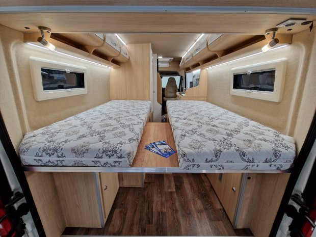 Wnętrze kampera z dwoma pojedynczymi łóżkami i drewnianą szafką między nimi.