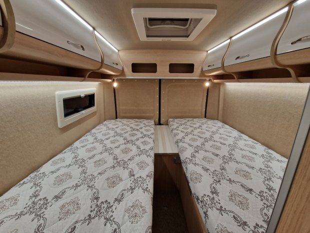 Wnętrze kampera: dwa łóżka pojedyncze z szafkami i oświetleniem nad nimi.