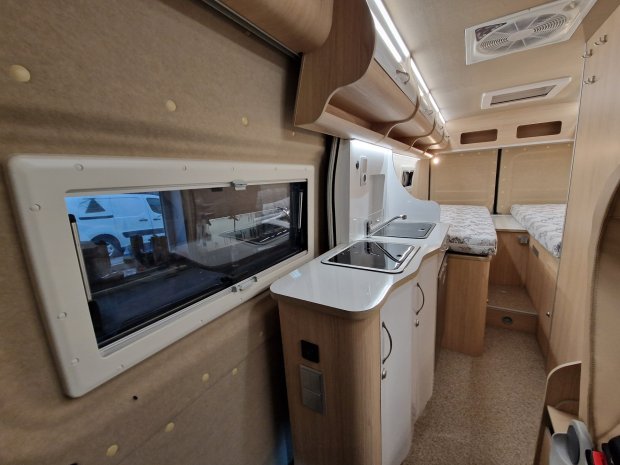 Wnętrze kampera: mała kuchnia ze zlewem i płytą, drewniane szafki i łóżka w tle