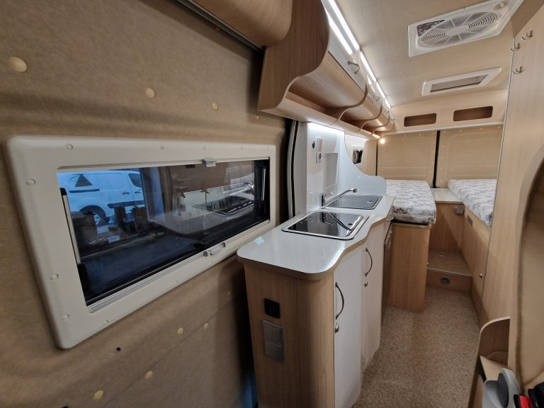 Wnętrze kampera: mała kuchnia ze zlewem i płytą, drewniane szafki i łóżka w tle