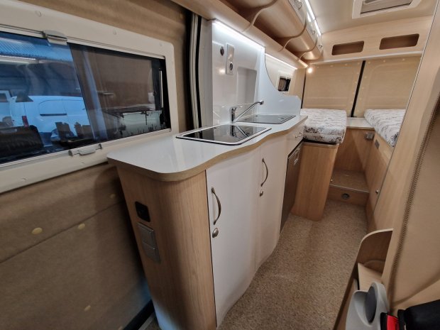 Wnętrze kampera z jasną kuchnią, zlewem, szafkami i dwuosobowym łóżkiem.