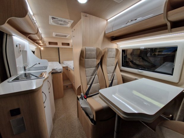Wnętrze kampera z aneksem kuchennym, stołem, dwoma fotelami i łóżkiem z tyłu.