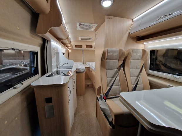 Wnętrze kampera z kuchnią, stolikiem, dwoma fotelami i łóżkiem z tyłu.