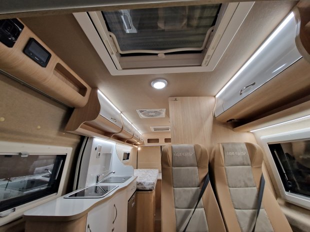 Wnętrze kampera z jasnymi szafkami, zlewem, dwiema fotelami i świetlikiem sufitowym.