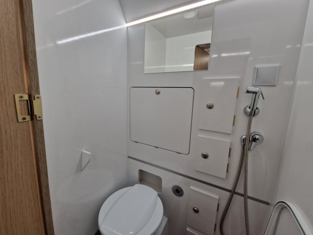 Kompaktowa biała łazienka z toaletą, prysznicem ręcznym, lustrem i schowkami.